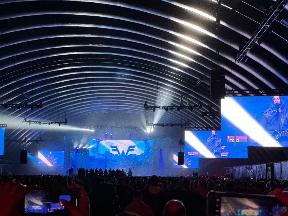 Shot of the RePlay MainStage far in the distance over the heads of a huge crowd of AWS nerds, with the Weezer logo on the center giant screen and video displays showing the band on the sides. The whole shot is inside an aircraft hanger style temporary building.