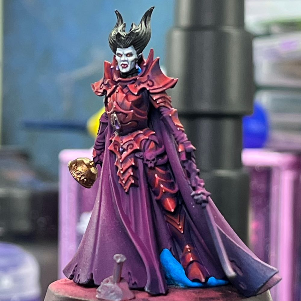 A female vampire lord in crimson armour, with rich details, and amethyst gems. In her right hand, she carries a chalice. In her left hand, a curved sword decorated with a rose.

The miniature is not fully painted yet. 