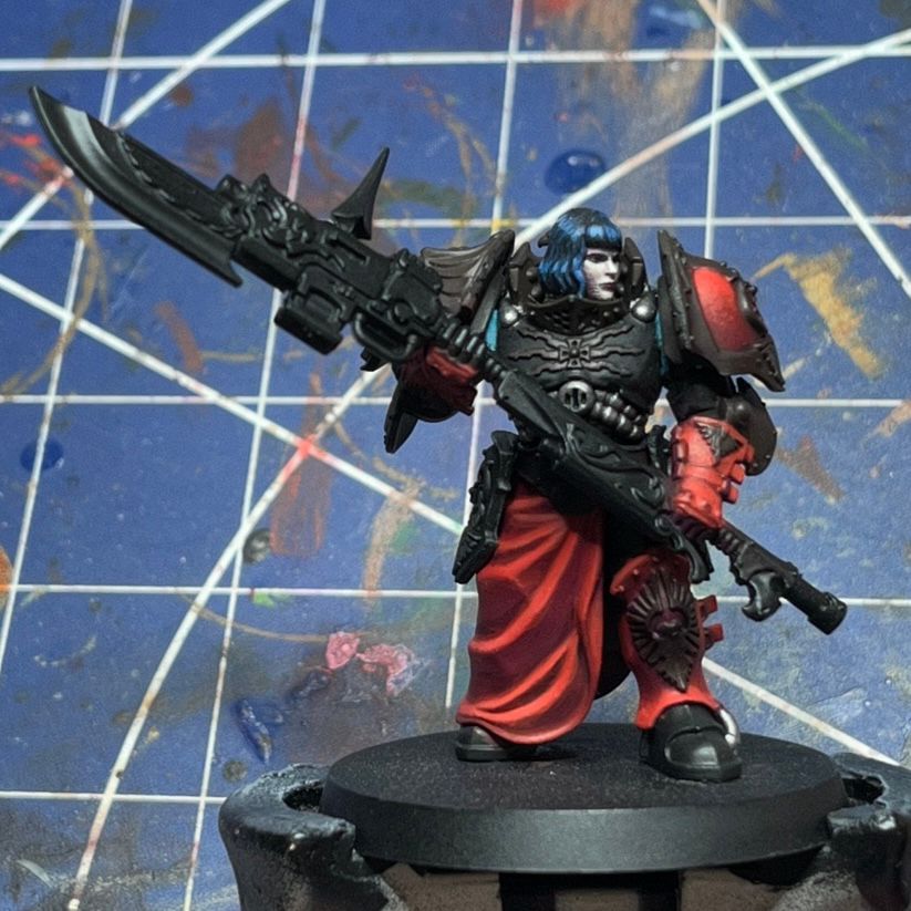 Picture of a miniature whose painting is still in progress. It’s an Adeptus Custodes Warden. She has bright blue hair and a pale complexion. Her armour is dark with red textiles.