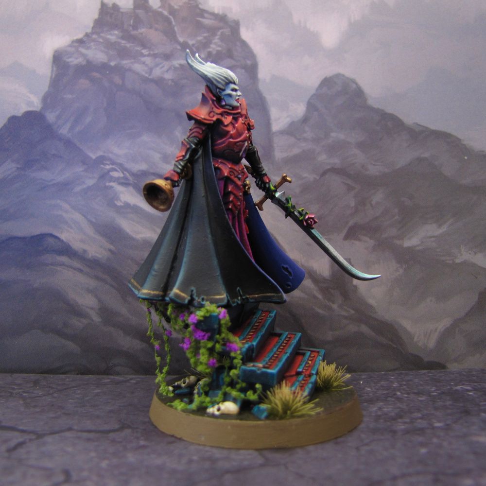 Miniature of a female Vampire Lord. She has cold white skin and strong features. He snow white hair is shaped like horns or bat ears above her head, and fiery, piercing eyes. Her lips are parted to show her fangs. She is covered from head to toe in crimson armour, with some gold studs and skull, as well as amethyst gems. A wine-coloured skirt flows from beneath her chest piece. Her cape is night blue on the outside, and dark violet on the inside. In her left hand, she holds an enchanted curved sword, with a blue-tinted blade, decorated with a red rose. The pommel and guard are gold. In her right hand, she carries an empty chalice, ready to be filled with the blood of her prey.

The lady is standing atop a flight of broken stairs, made of dark teal stone and blood red steps with inlaid golden teeth pattern. Vines with purple flowers are growing along one side of the steps. Broken masonry and bones are scattered around the damp ground amid the grass and soil.