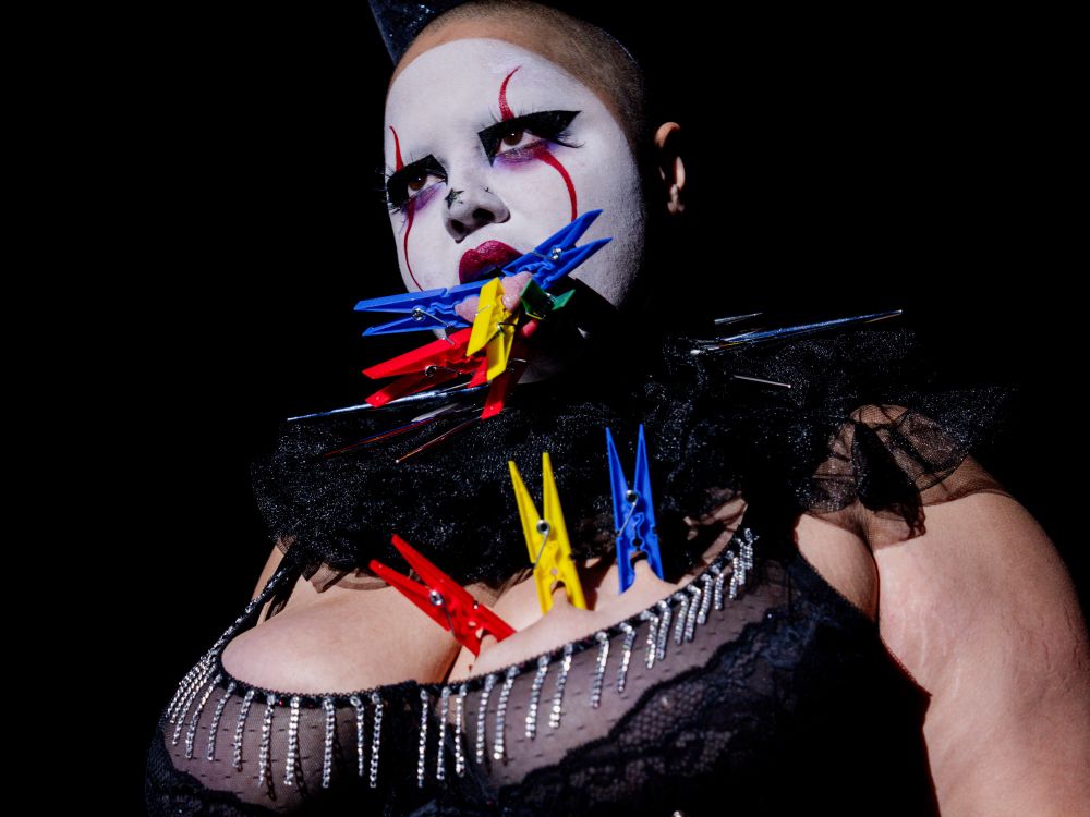 A white face clown with 9 colorful clothes pins clipped to their tongue, bottom lip, and right breast