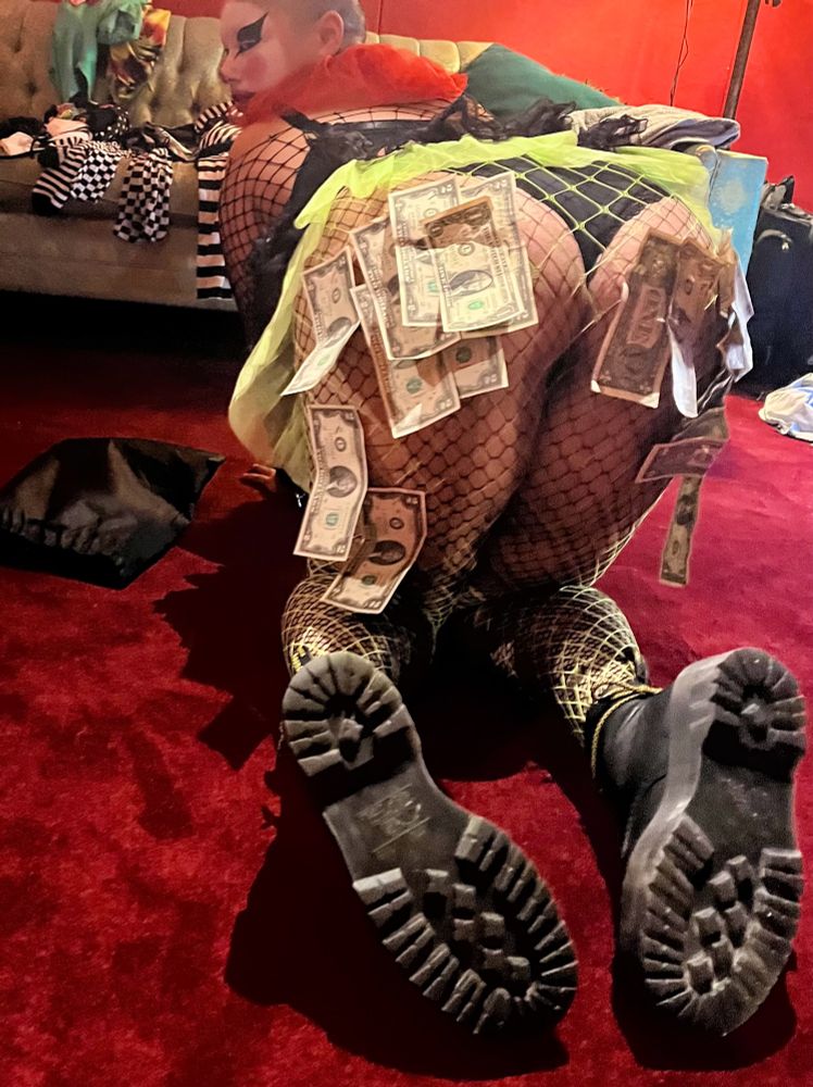A clown in fishnets on all fours on a red carpet, in front of a light green couch. Their butt and thighs covered in money, mostly $2 bills