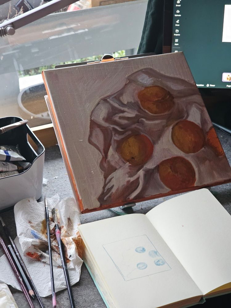 A picture of a small canvas resting on a table with a sketchbook depicting 5 peaches on a white table cloth. Next to the sketchbook are brushes and a tissue.