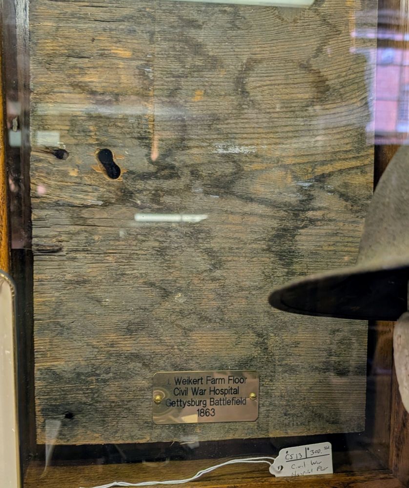 A piece of wooden flooring in a case. There is a small plaque on the wood that says "Weikert Farm Floor, Civil War Hospital, Gettysburg Battlefield, 1863". The floor is stained with old bloodstains all across it ans there are two small holes in it. There is a portion of a world war one helmet on a Styrofoam head slightly on front of the display. The Weikert family's buildings were indeed used during the American Civil War. At least six men died right in the family's parlor, and severed body parts from emergency amputations were piled outside of the windows. Later, the family's missing rug was found to have been used to line a mass burial pit for dead soldiers. 