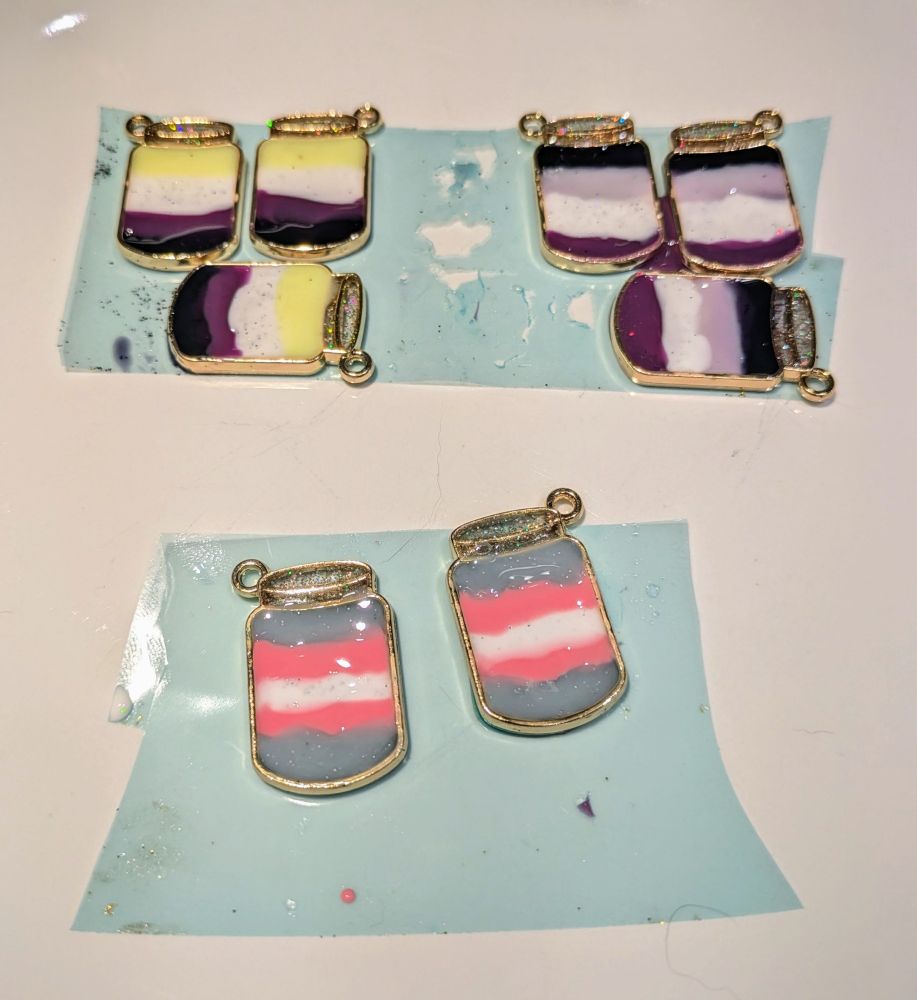Several necklace pendants shaped like closed mason jars. Inside each jar are various pride colors. Three are non-binary colors: yellow, white, purple, black. Three are asexual colors: black, gray, white, purple. Two are trans colors: blue, pink, and white. All of the pieces are still on resin tape on top of a white plate.