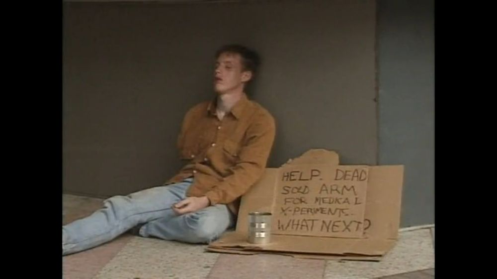a man lays slumped against a wall. he has a cardboard sign that reads, "help, dead. sold arm for medical x-periment. what next?"