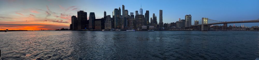 Sun sets in the left of the frame with the Lower Manhattan skyline in the center and the Brooklyn Bridge to the right. The East River forms the foreground of the picture. 