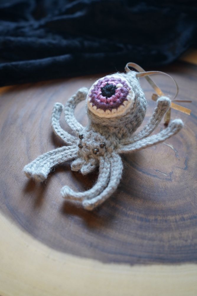 A gray crochet spider with a purple eyeball as its abdomen sits on a wooden stump slab with a black velvet cloth in the background. The spider has a shimmery gold ribbon tied to its abdomen. 