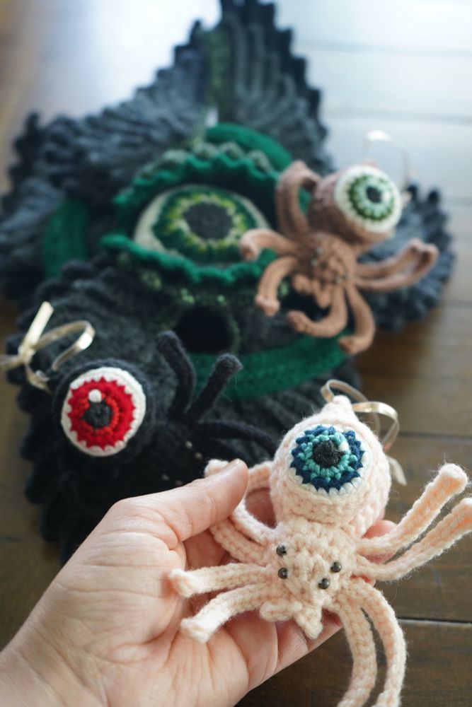 There are multiple crochet pieces resting on a wooden table. A pink spider with a blue eyeball for a body is being held by a hand. There is a black spider with a red eyeball body, a brown spider with a green eyeball body, and a black and grey seraph with a large green eyeball center in the background. 