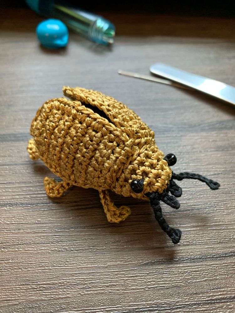 A small, golden crochet beetle sits on a wooden table. 