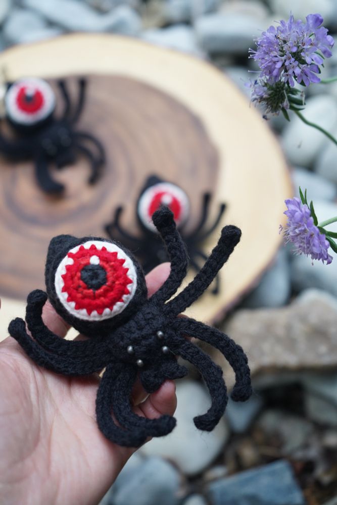 A hand holding a black crochet spider. The spider has a red eyeball for an abdomen and has silver eyes. There are two more crochet spiders in the background just like it resting on a wooden stump slab. There is also a light purple pincushion flower within frame. 