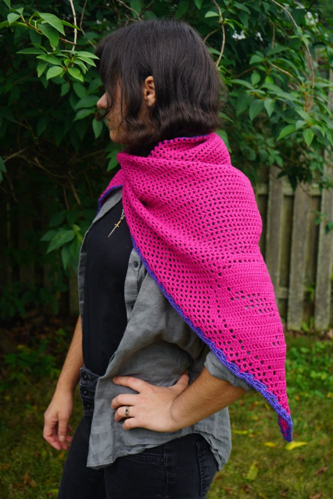 A person with dark brown hair covering their eyes and olive toned skin stands with one hand on their hip looking into the distance in front of a lush green forsythia bush. They are wearing a black shirt, grey over shirt, black pants, and a handmade crochet magenta shawl with a purple border. 