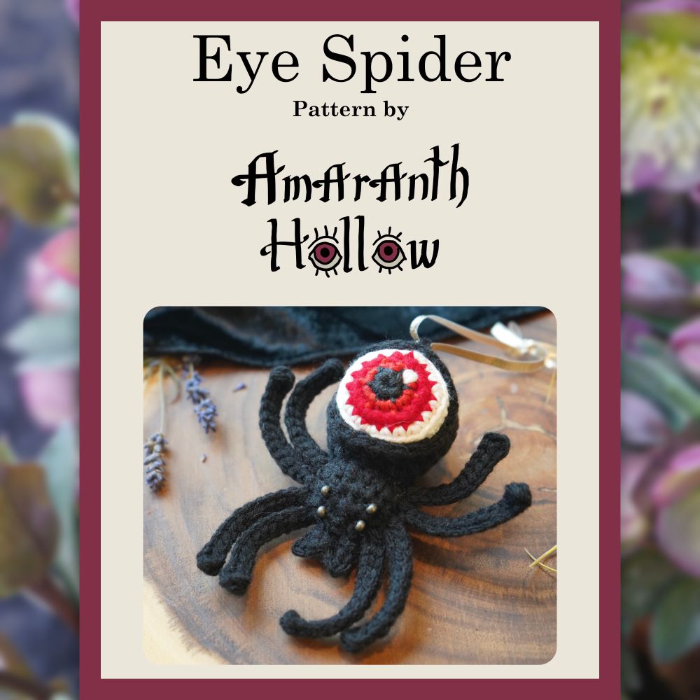 A picture of a black crochet spider with a red eyeball for an abdomen framed by a beige and magenta border. Within the frame is the text “Eye Spider Pattern by Amaranth Hollow.” Outside the frame is a blurred photo of hellebore flowers as the background. 