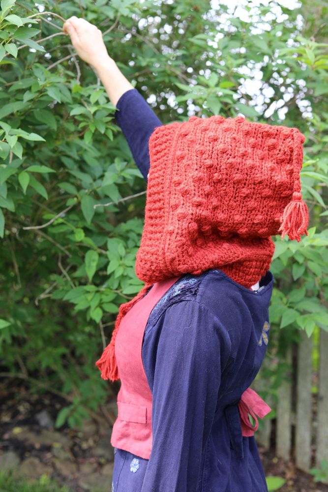 Someone in a blue dress reaching up a forsythia bush wearing a reddish orange handmade crochet hood. The hood is textured with bobbles and tassels. 