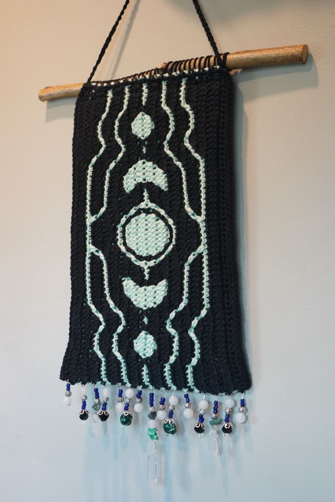 A black and mint green crochet tapestry hangs from a stick dowel in front of a neutral toned wall. The mint part of the tapestry is a geometric design similar to the moon phases. At the bottom of the tapestry is a fringe made of small blue glass beads, quartz crystals, and black beads. 