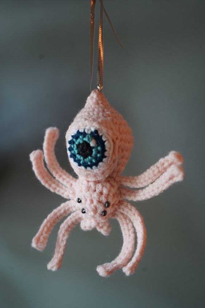 A peachy pink crochet spider with a large blue eyeball attached to its abdomen hangs from a golden ribbon in front of a bluish green background. 
