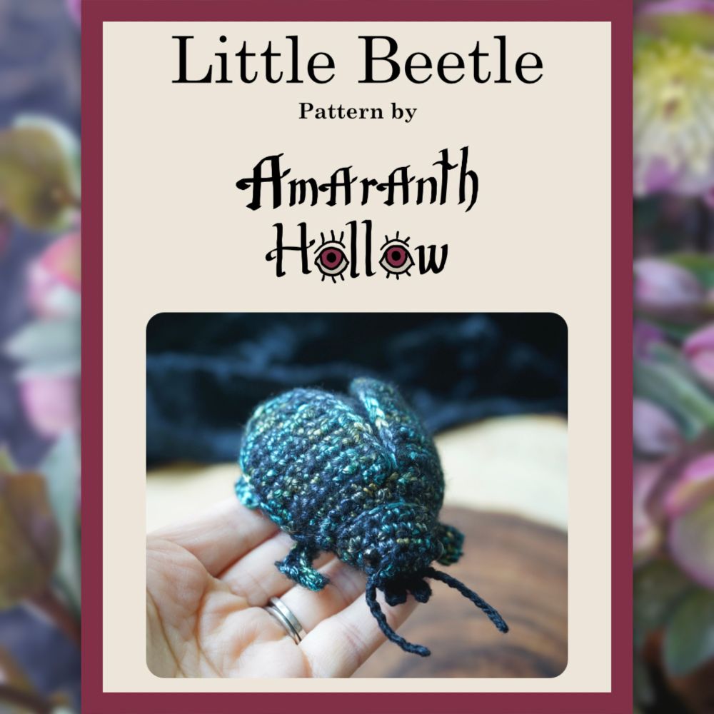 Photo text says “Little Beetly Pattern by Amarabth Hollow” on a cream background and shows someone holding a blue crochet beetle. The border of the photo is a blurred picture of hellebore flowers in a garden. 