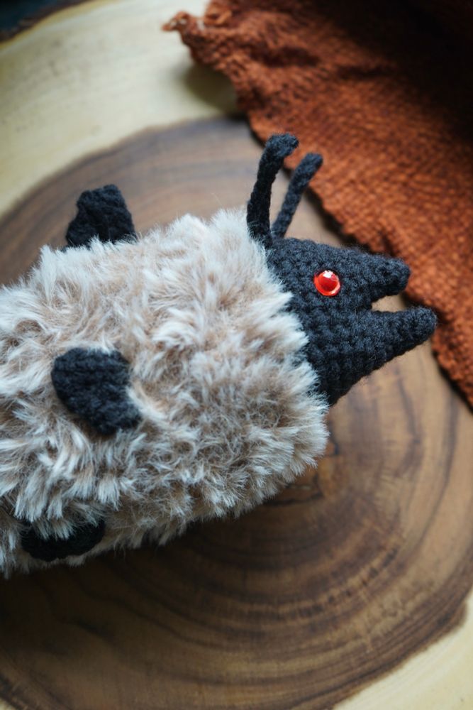 A black crochet fish with red eyes and a tan, fuzzy body rests on a wooden stump.