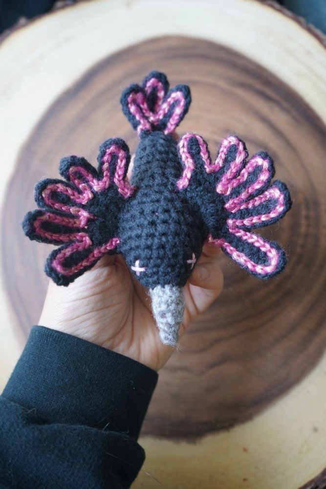 A hand holds a black crochet crow above a wooden stump platform. The crow has light pink “X” eyes, a grey beak, and its wings and tail are outlined in shimmery pink yarn. 