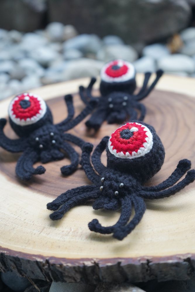 Three crochet spiders rest on top of a wooden stump slab nestled amongst large pebbles on the ground. The three spiders are black with grey metallic beaded eyes. Each spider has an eyeball for an abdomen with a red iris. 