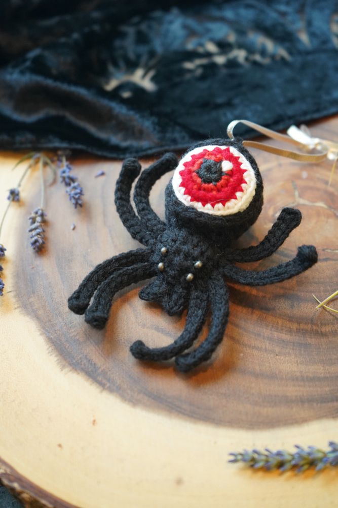 A black crochet spider with a red eyeball as its abdomen is resting on a stump-like wooden platform scattered with lavender flowers.