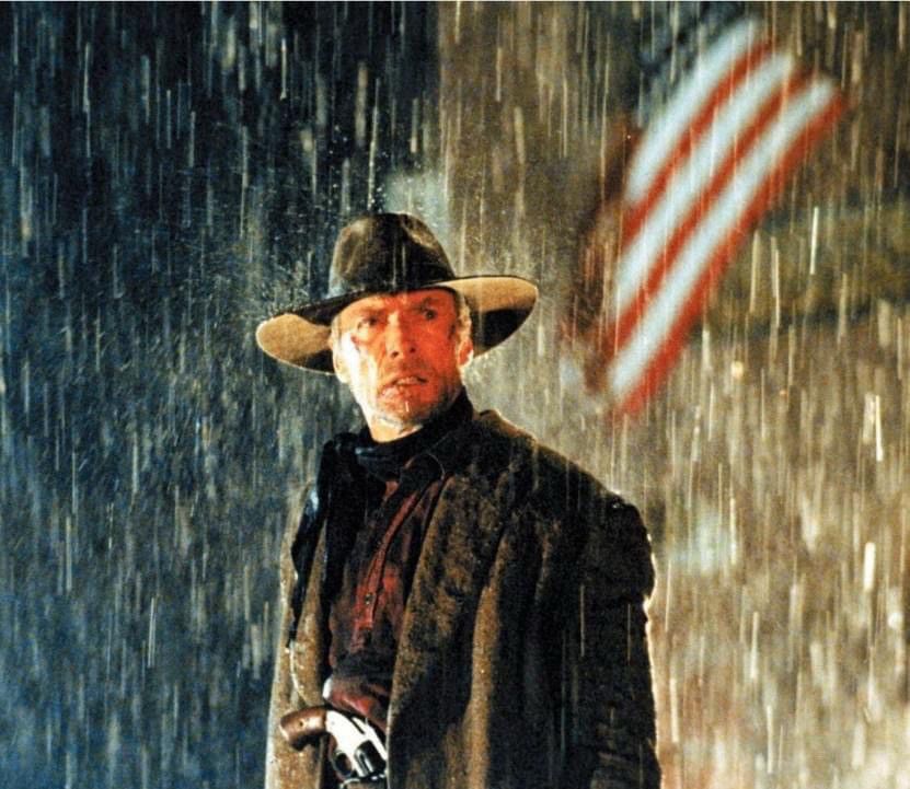 Clint Eastwood as William Munny at the end of UNFORGIVEN, standing in the rain at night beneath an American flag