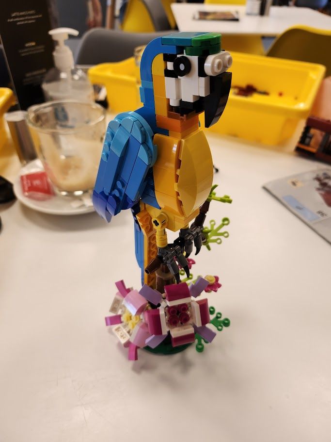 A Macaw and some flowers made of lego on a cafe table. An empty coffee cup sits in the background