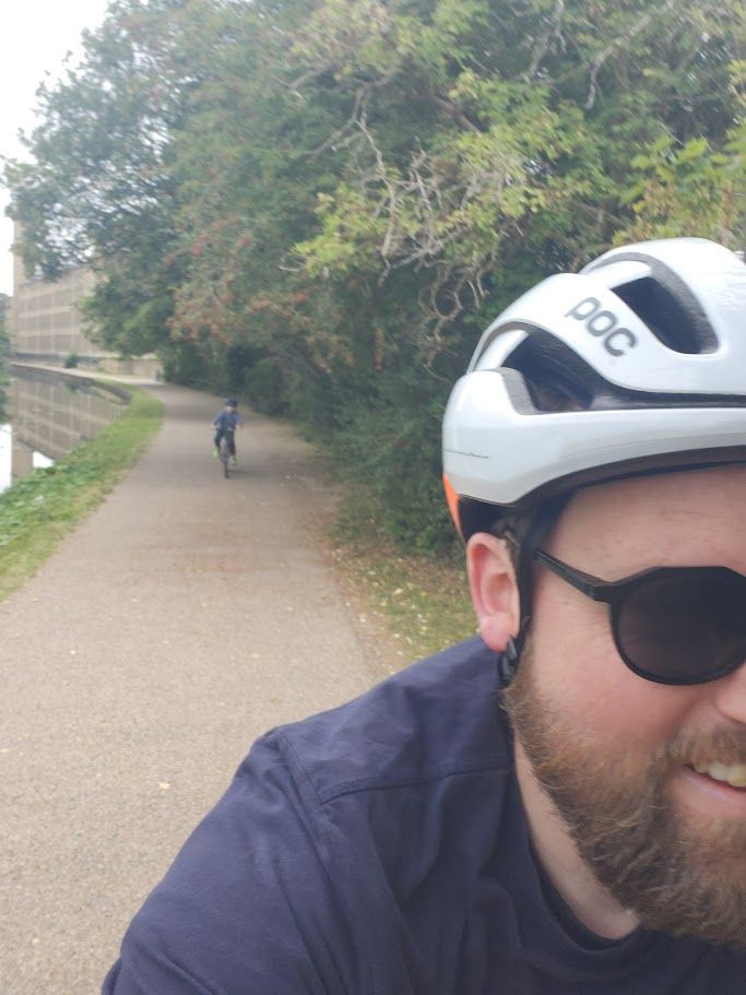 Selfie view of a man in a white bicycle helmet with a canal toe path in the background. A child rides along in the distance