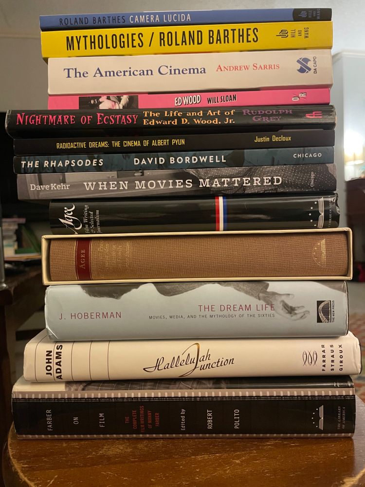A stack of books, which include, from top to bottom:

-Camera Lucida: Reflections on Photography, by Roland Barthes

-Mythologies, by Roland Barthes

-The American Cinema, by Andrew Sarris

-Ed Wood, Made in Hollywood USA by Will Sloan

-Nightmare of Ecstasy: The Life and Art of Edward D. Wood Jr., by Rudolph Grey

-Radioactive Dreams: The Cinema of Albert Pyun, by Justin Decloux

-The Rhapsodes: How 1940s Critics Changed American Film Culture, by David Bordwell

-When Movies Mattered: Reviews from a Transformative Decade, by Dave Kehr

-Two hardcover James Agee collections from Library of America

-The Dream Life: Movies, Media, and the Mythology of the Sixties, by J. Hoberman

-Hallelujah Junction, by John Adams

-Farber on Film: The Complete Film Writings of Manny Farber