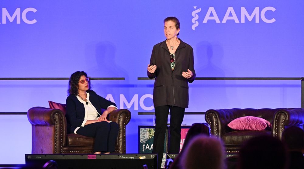 Two speakers on stage at an AAMC conference; one is seated and the other is standing, gesturing while speaking. They are in front of a backdrop with the AAMC logo.