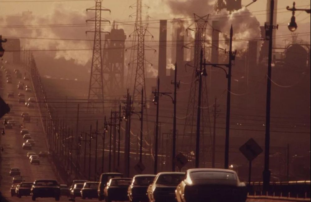 A vintage photo from the National Archives shows cars driving down a road as smoke spews from factories in the distance and air pollution hangs over everything.