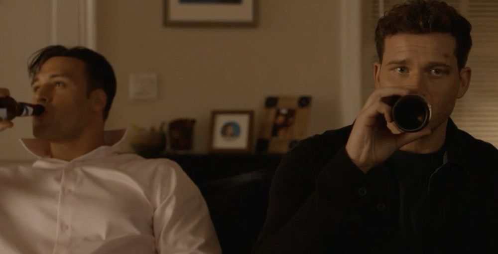 Ryan Guzman as Eddie Diaz in 9-1-1 on ABC wearing a light pink button-down shirt with the collar popped as he leans back on a couch, looking content. Beside him, Oliver Stark as Evan Buckley looks dejected as he sits forward on the same couch and drinks his own beer. 