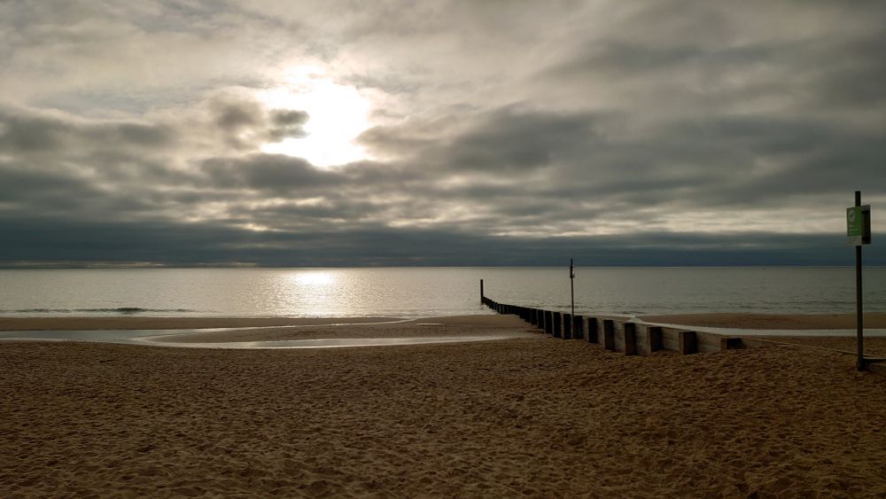 A colour photo showing the view across the beach to the sea early Sunday monring. In the foreground, the beach is empty, but covered in footprints and other traces of mostly human and canine activity. The sea, across the middle of the photo, looks somewhat bracing. The cloudy sky is hiding the sun, whose presence is indicated by a bright white splodge just left of centre.