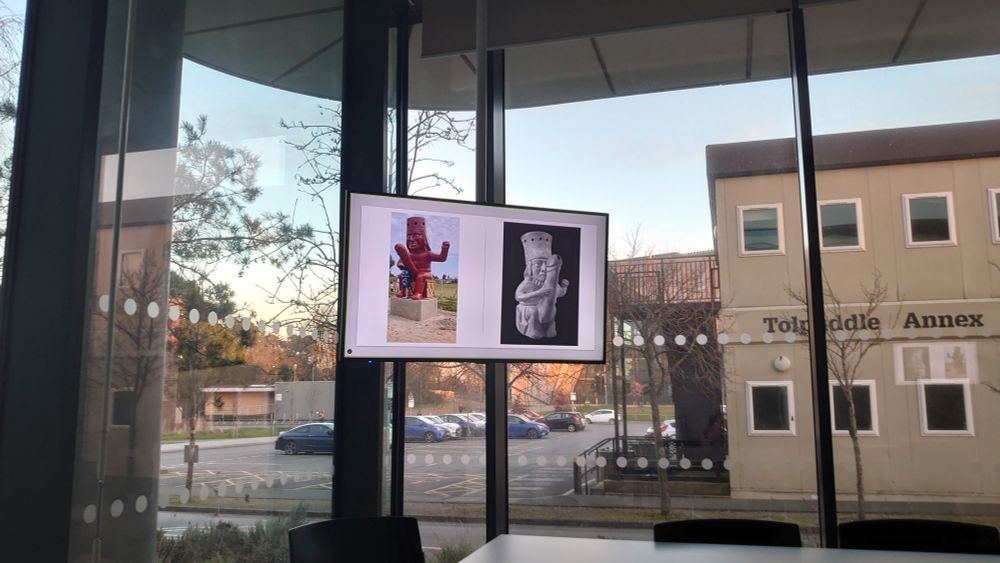 A colour photo taken inside one of the lecture rooms in the Fusion Building at Bournemouth University, capturing the view out of the rather high windows of the car park and neighbouring buildings. In the centre, inside the room, is a screen containing two images of figures best described as priapic.