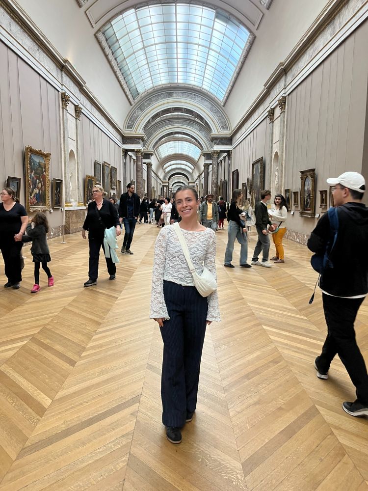 Girl stood in the centre of a gallery hall wearing a white top and navy trousers. 