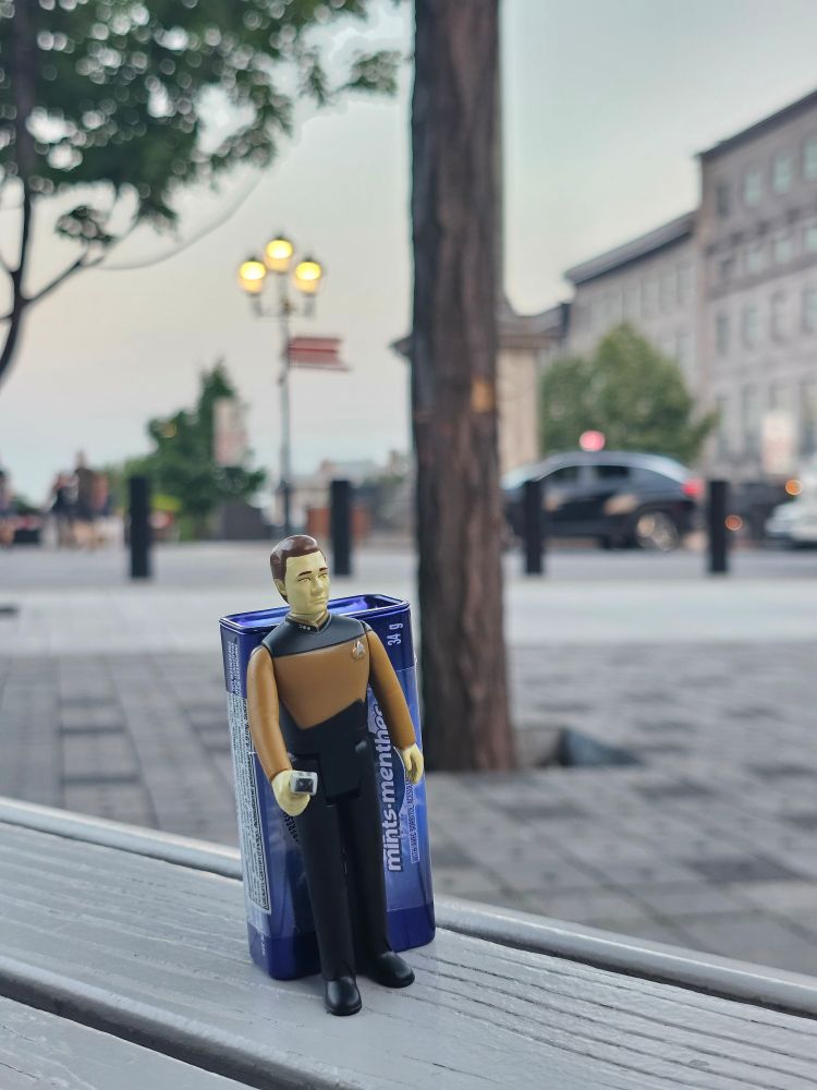 A picture of Data toy action figure holding the phaser. Behind him is a case of mint candy (I had to put it there so that he won't fall 😭). He is standing on gray bench. The background is that a bunch of buildings standing on the right side, one streetlight, some scattered trees, one black/blue (????) car and the floor is made of stone (idk what to describe lol). It's all blurred, though