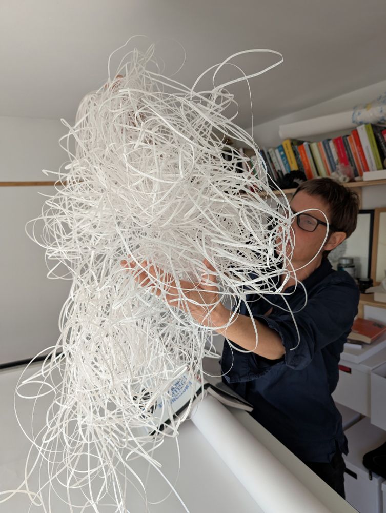 A woman wearing glasses and a dark shirt holds a large pile of very thin, curly strips of paper. The pile is shaped like a cloud. She is holding the pile over a white table with a roll of paper on one side and a book opened face-up resting on the roll. Behind her are bookshelves with books on them.