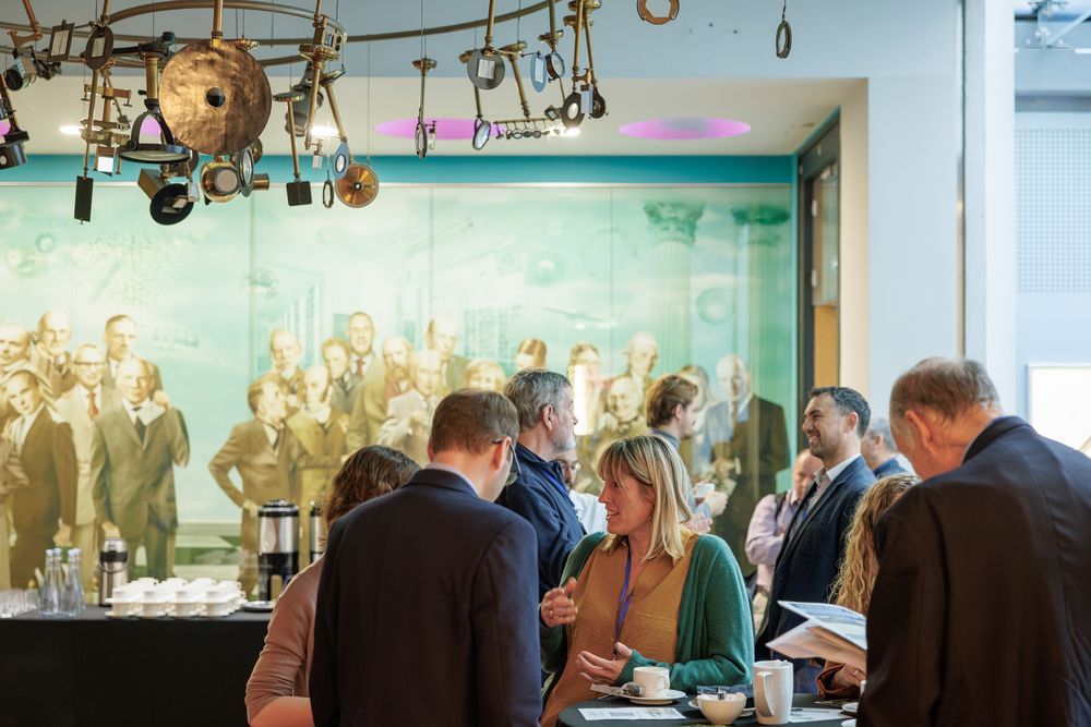 Groups of people stand talking to each other and smiling. On the back wall is a large print of people wearing suits. Above the people talking is a chandelier made of scientific instruments. A table with coffee urns, cups, water bottles and glasses is in front of the wall with the large print. 