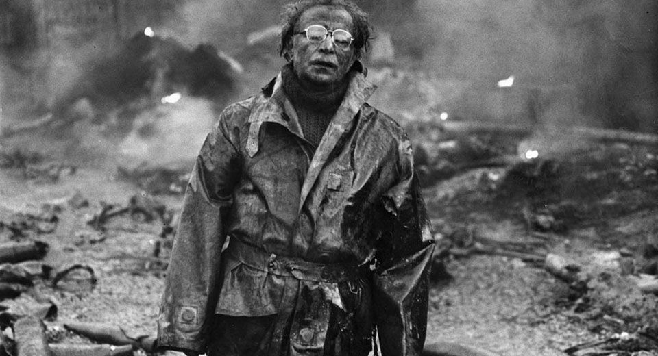 A wide shot of a man with glasses and a coat, looking up towards the sky over the camera's pov with chaotic & rubble behind him