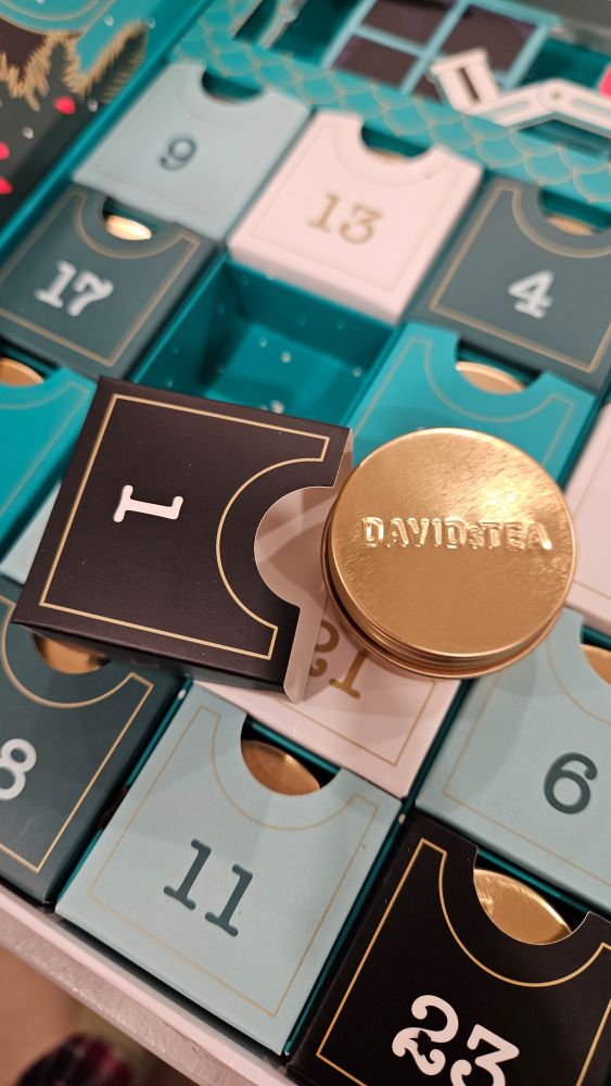 uma latinha de chá com a inscrição "Davids Tea" ao lado de uma caixinha de papel com o número 1, sobre uma caixa de calendário do advento onde estão várias outras caixinhas numeradas