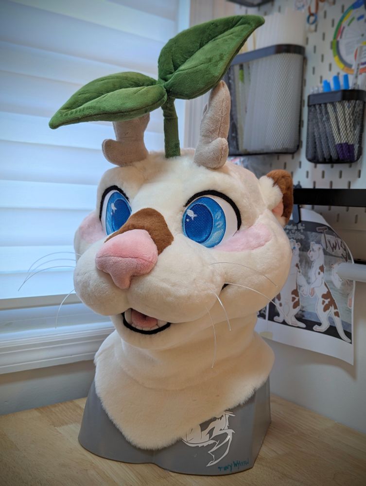 Otter fursuit head that is cream colored with a brown spot on nose and ear, along with a leaf on top of head and small horns.