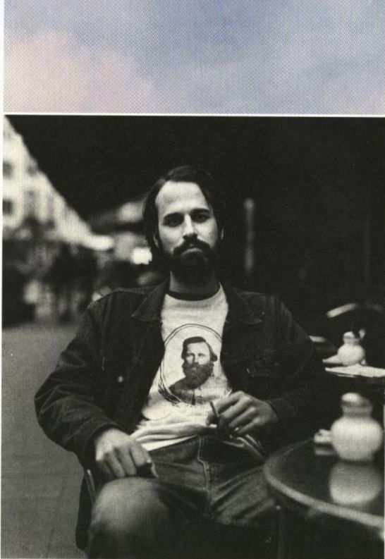 A black and white photo of David Berman, a skinny man with dark hair and beard, holding a cigarette while sitting at a cafe table.