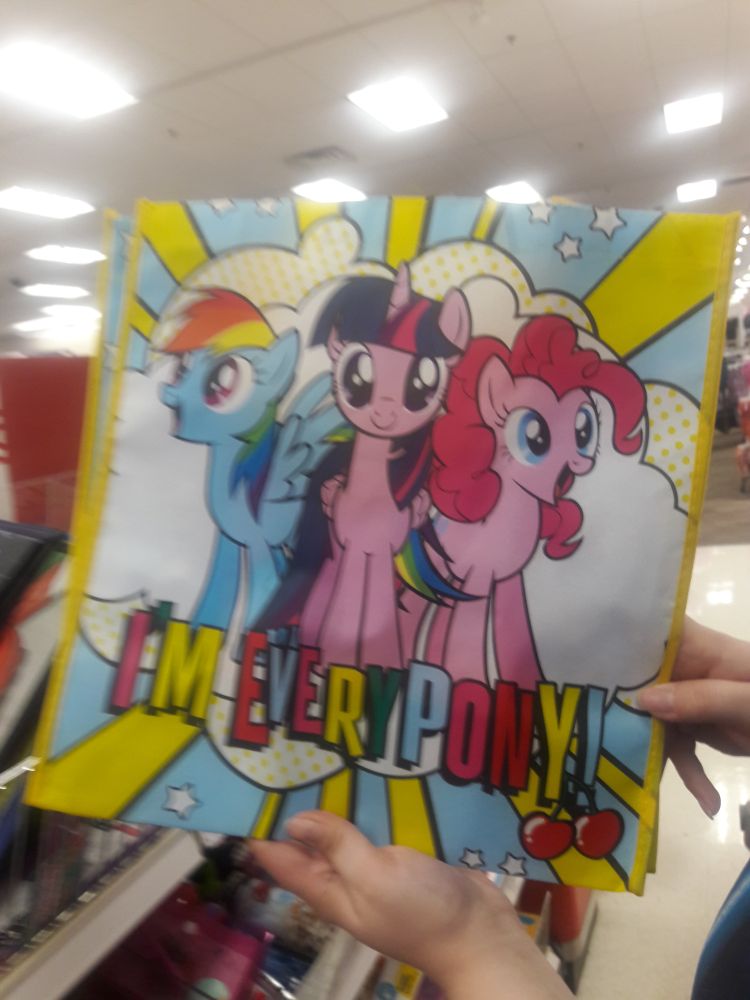 Photo of a My Little Pony Gen 4 tote bag from walmart, displaying Twilight Sparkle in the center with Rainbow Dash and Pinkie Pie on either side of her smiling excitedly. There's an explosion of clouds behind them. There is text below them in disorganized rainbow colors that has the confusing english phrase "I'm Everypony!" with two cherries hanging off the end.
