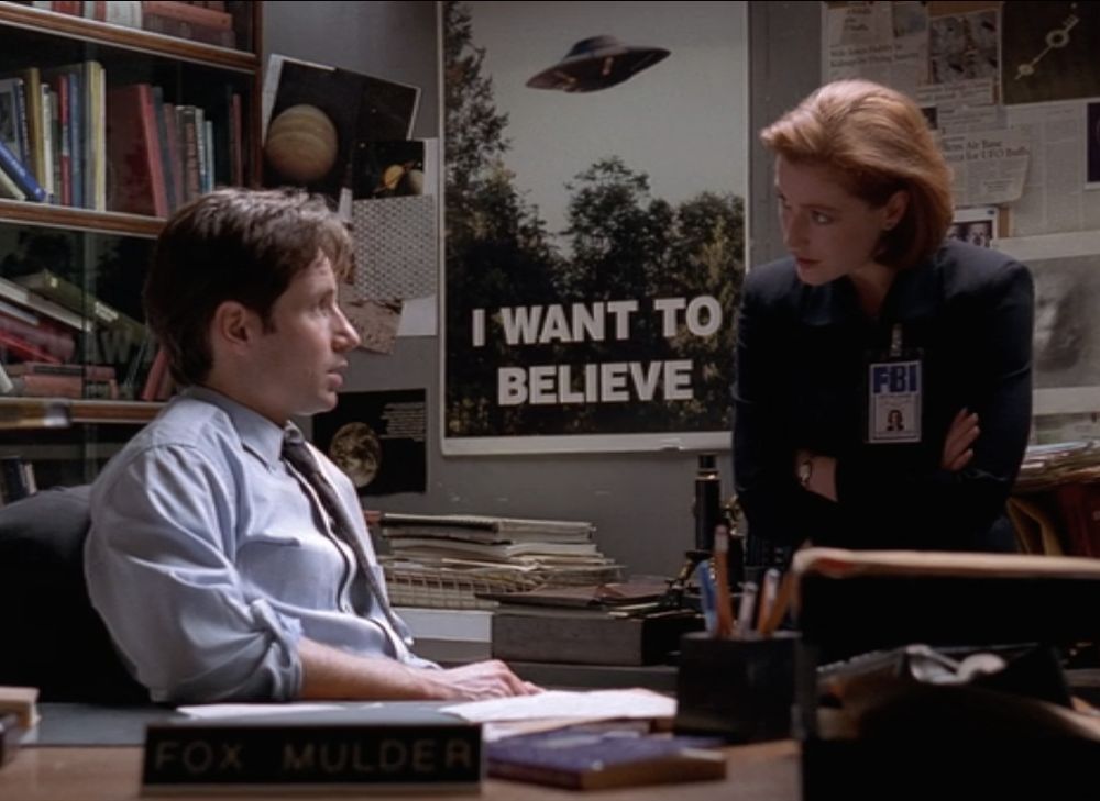In Mulder's shambolic cubicle, Mulder and Scully debate the existence of the paranormal. On the wall behind them, a poster featuring a grainy UFO photo and the heading "I WANT TO BELIEVE"