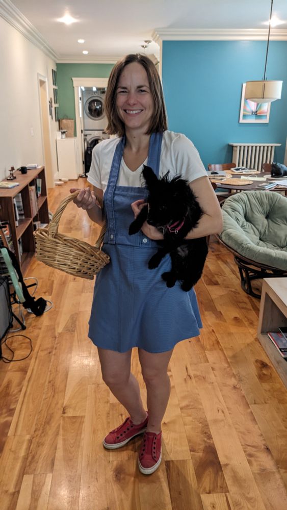 A woman dressed as Dorothy from the wizard of oz holding a basket and small black dog