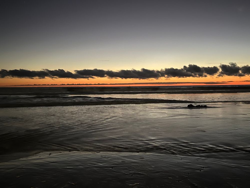 Orange sky with a line of dark grey clouds just above the horizon line of the ocean. Low shimmering water in the foreground. 
