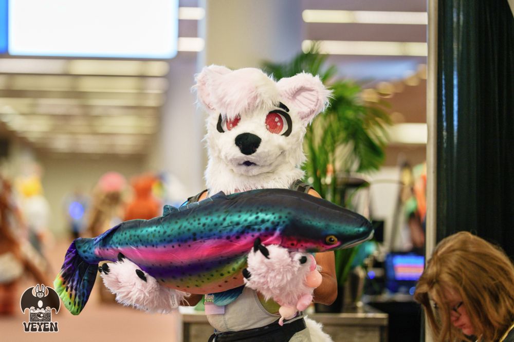A fursuiter is holding a prop fish at Anthrocon 2025.