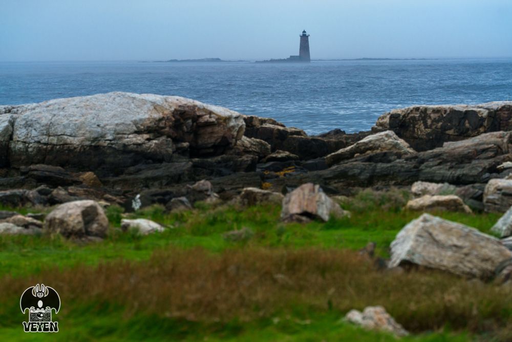 It’s a photo of a landscape. There’s a lighthouse in the distance, surrounded by ocean. The water and sky are dark. 
