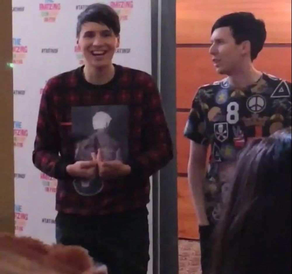 Photo of Dan and Phil standing in front of their meet and greet backdrop at their second TATINOF Glasgow show. Dan is laughing and speaking out to the crowd of fans while Phil is turned towards Dan and looks to be in the middle of saying something both to Dan and to the phannies in front of them.