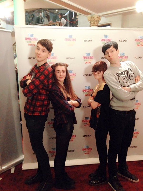 Photo of Dan and Phil at the TATINOF Folkestone meet and greet, posing back to back with phandom icons Katherine and Eden (writers and performers of the 2009, 2012, and 2022 songs). Everyone has their arms folded and are confidently staring the camera down.
